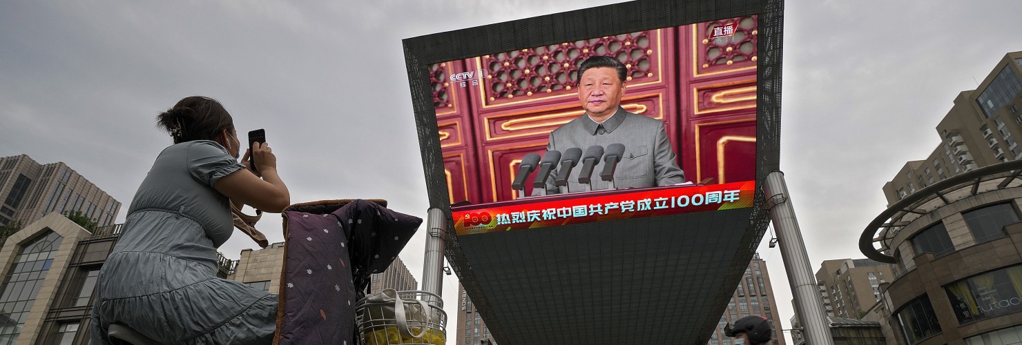 A woman on her electric-powered scooter films a large video screen outside a shopping mall showing Chinese President Xi Jinping speaking during an event to commemorate the 100th anniversary of China's Communist Party at Tiananmen Square in Beijing