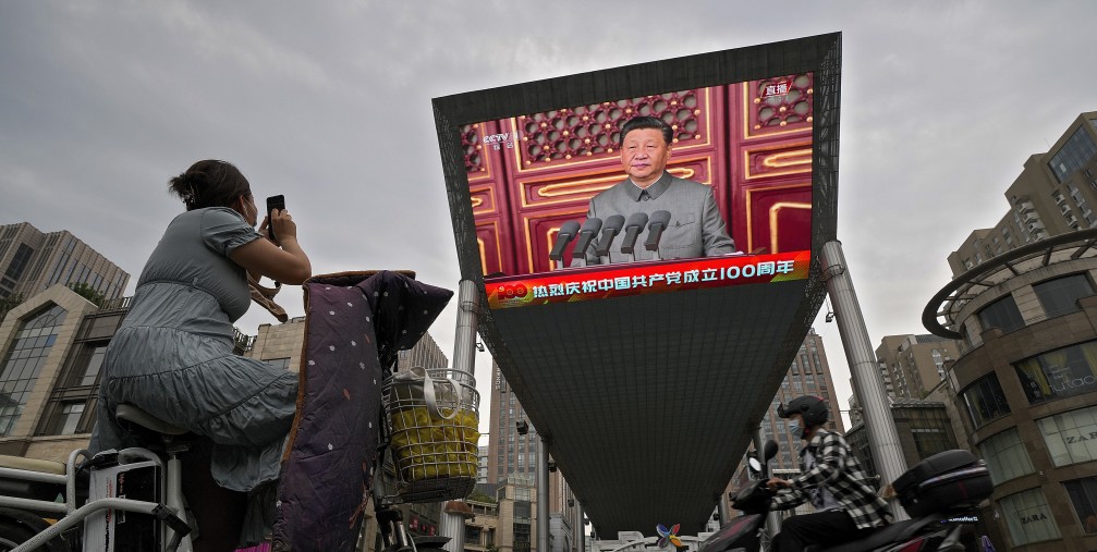 A woman on her electric-powered scooter films a large video screen outside a shopping mall showing Chinese President Xi Jinping speaking during an event to commemorate the 100th anniversary of China's Communist Party at Tiananmen Square in Beijing